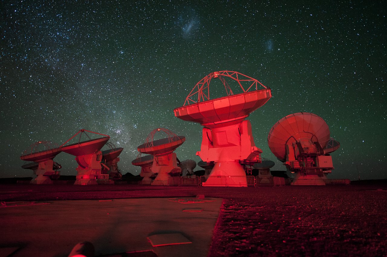 ALMA antennae bathed in red light. in the background there is the southern Milky Way on the left and the Magellanic Clouds at the top. Credit: ESO/C. Malin